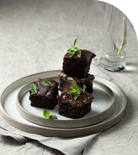 Gazdag csokoládés brownie-kockák fényes csokoládébevonattal és friss mentalevelekkel, elegáns kávéházi stílusban, minimalista kerámia tányéron tálalva.